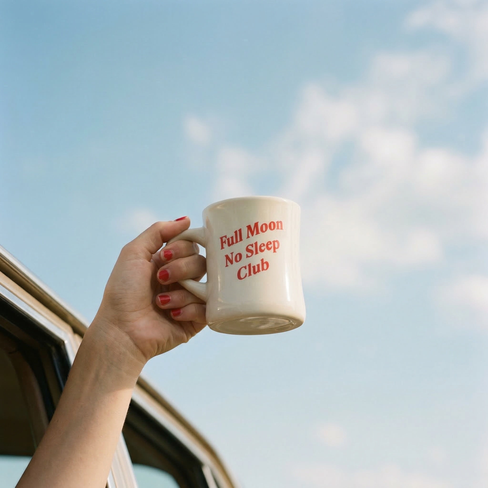 Hand holding a mug with 'Full Moon No Sleep Club' text against a blue sky.